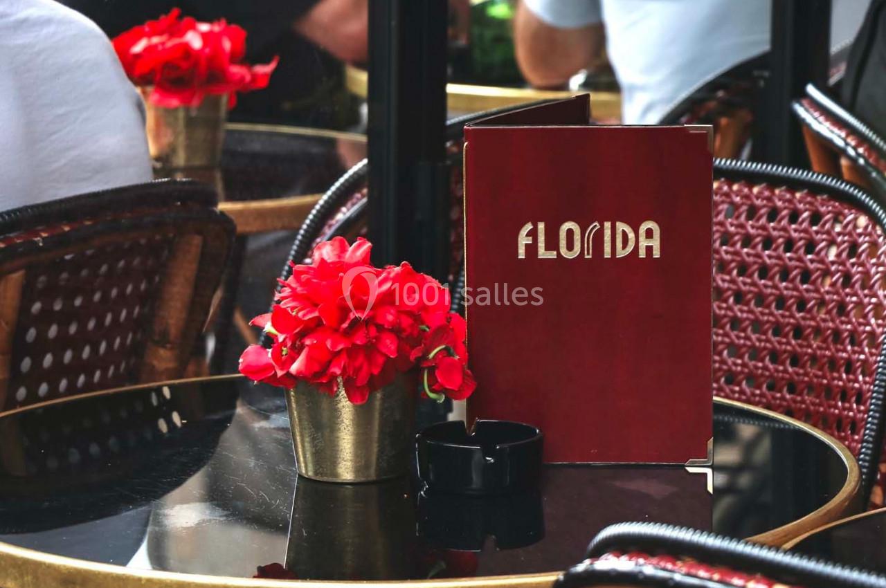 Des tables de café en terrasse avec des fleurs rouges et un menu rouge marqué ’Florida’ posé sur l'une d'elles.
