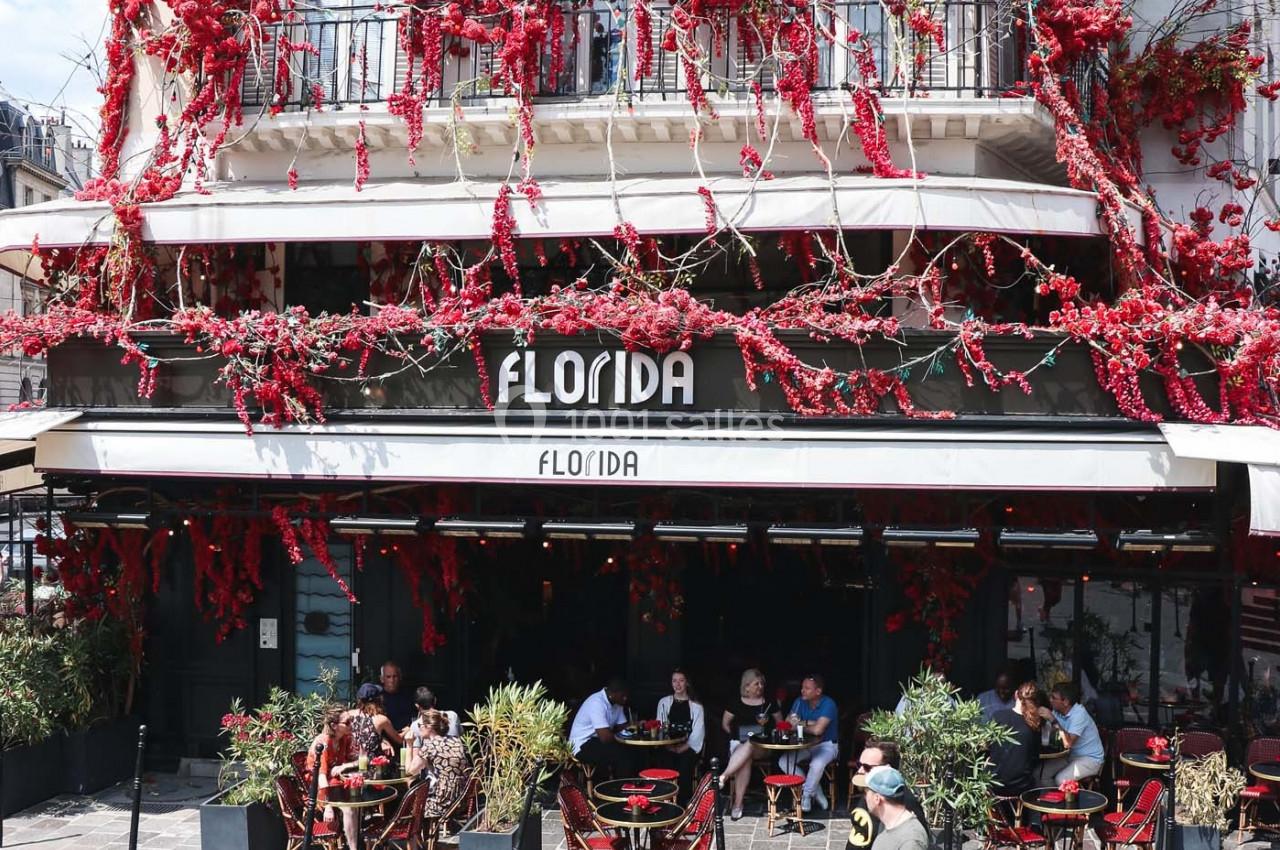 Façade d'un café avec terrasse, ornée de fleurs rouges, des passants marchent sur une rue pavée au premier plan.