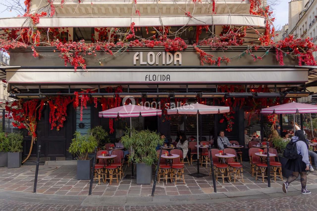 Façade d'un café avec terrasse, décorée de fleurs rouges, tables et chaises disposées sur un trottoir pavé.