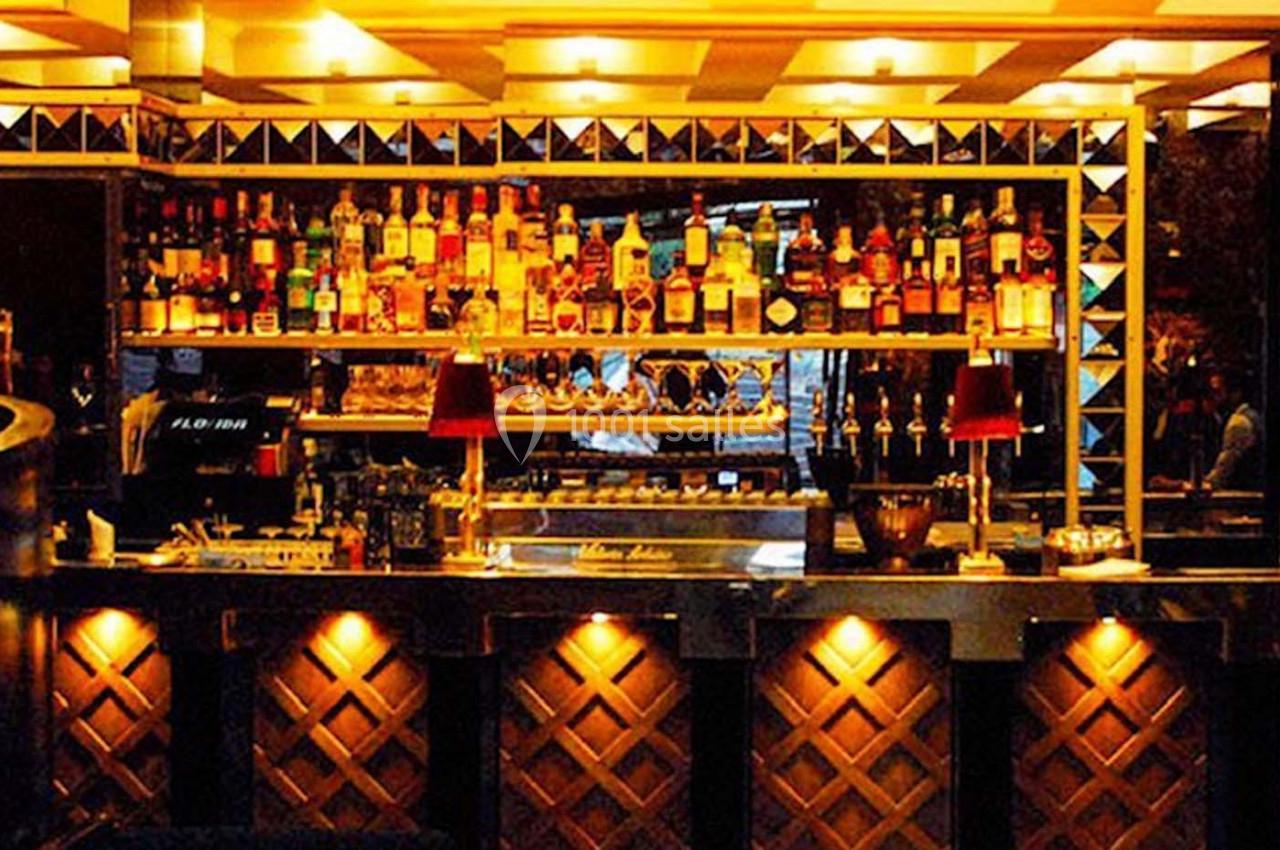 Bar éclairé avec des étagères remplies de bouteilles, des lampes rouges et un comptoir en bois sculpté.