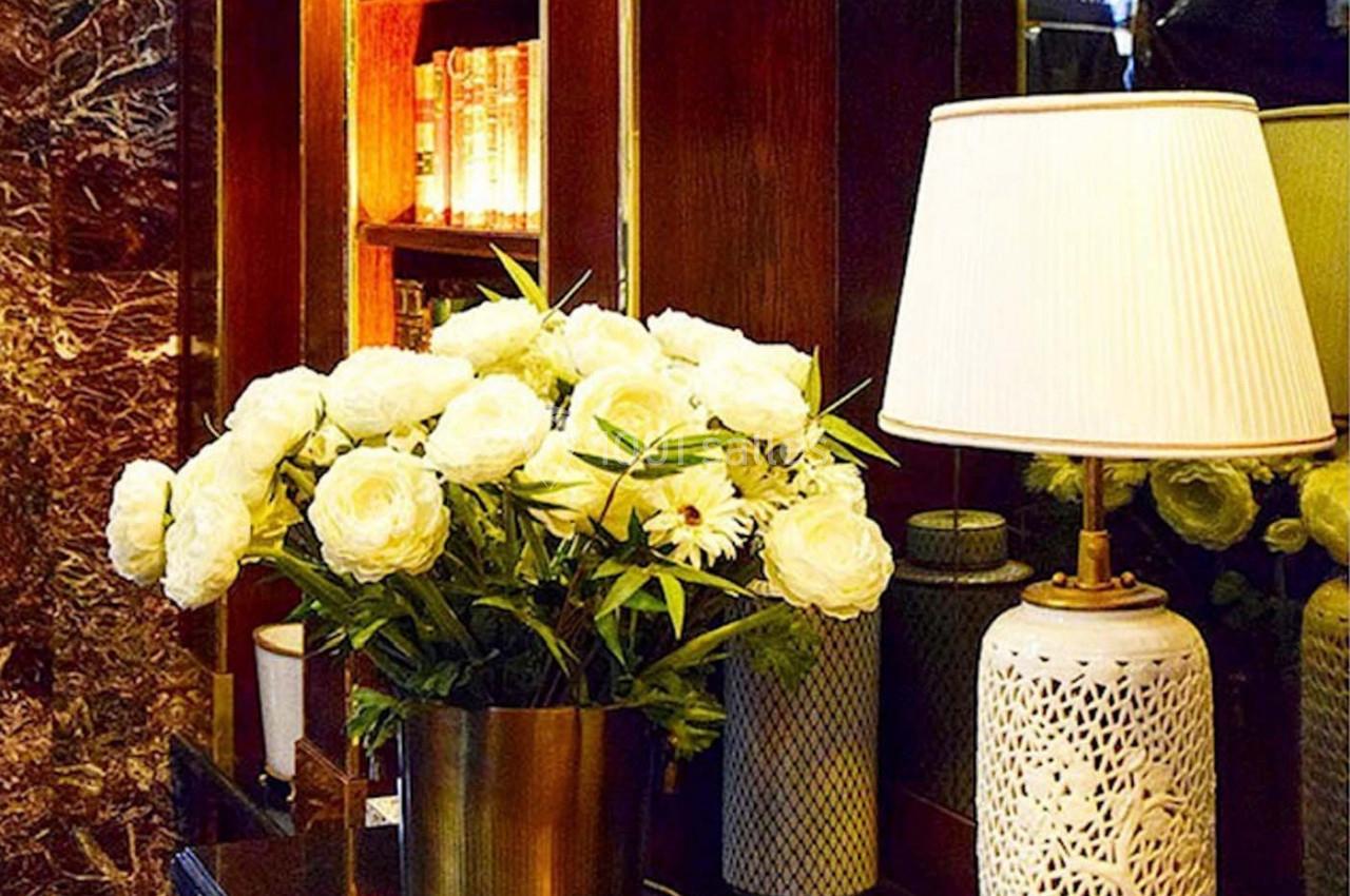 Bouquet de fleurs blanches dans un vase doré sur une table, avec une lampe et des étagères en arrière-plan.