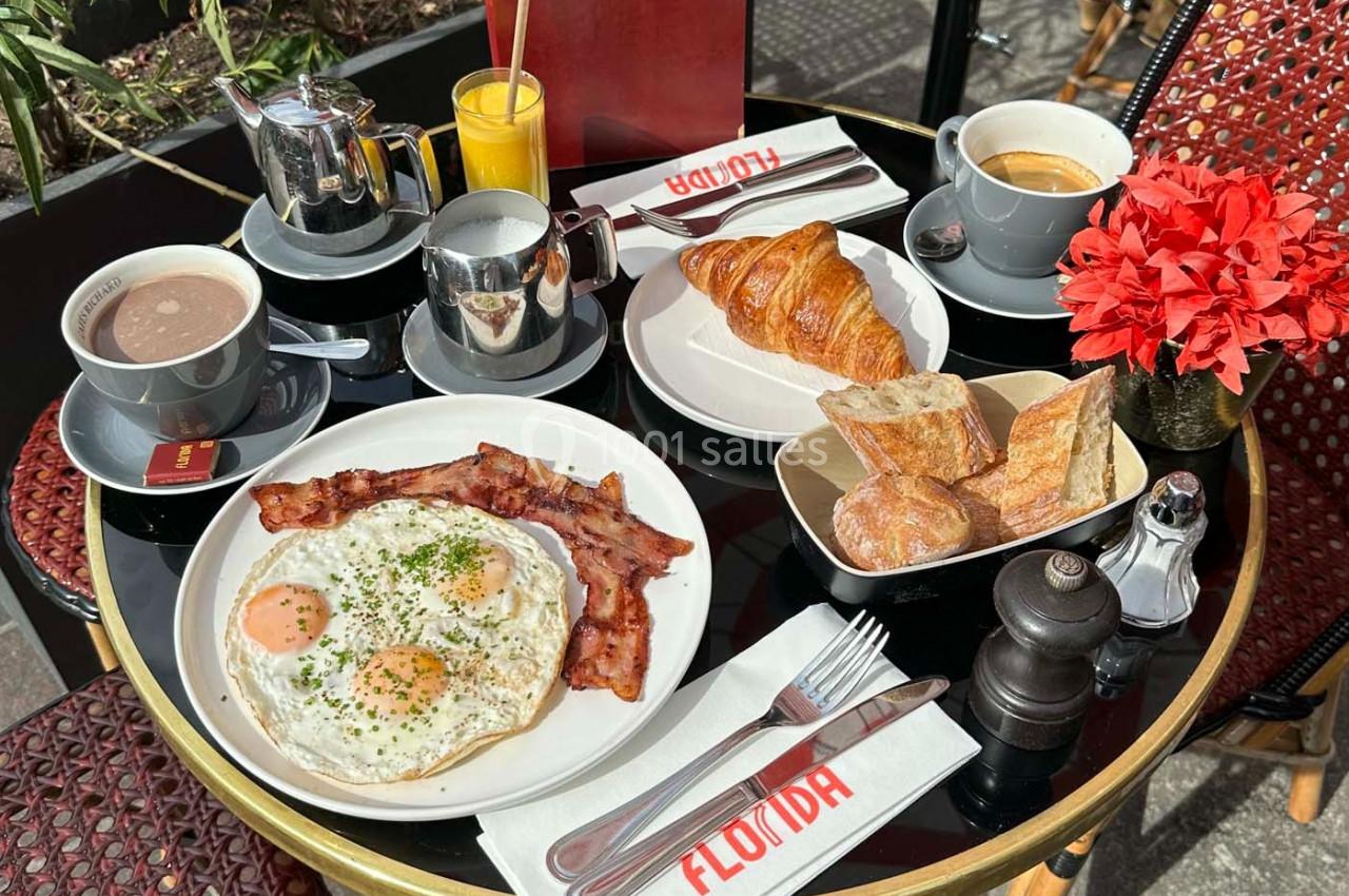 Petit-déjeuner en terrasse avec œufs, bacon, croissants, pain, boissons chaudes et jus d'orange sur une table ronde.