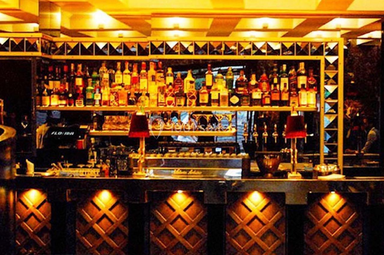 Bar éclairé avec des étagères remplies de bouteilles d'alcool, des lampes rouges et un comptoir en bois sculpté.