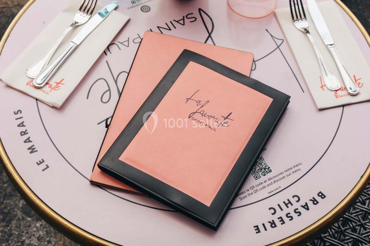 Deux menus roses posés sur une table ronde avec des couverts et des serviettes imprimées.