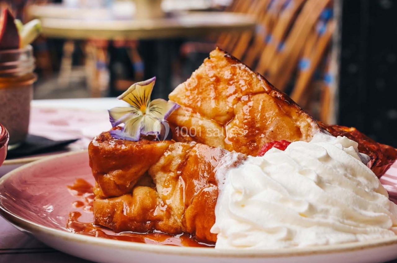 Tranche de pain perdu garnie de chantilly, fleur comestible et sirop, servie dans une assiette rose.