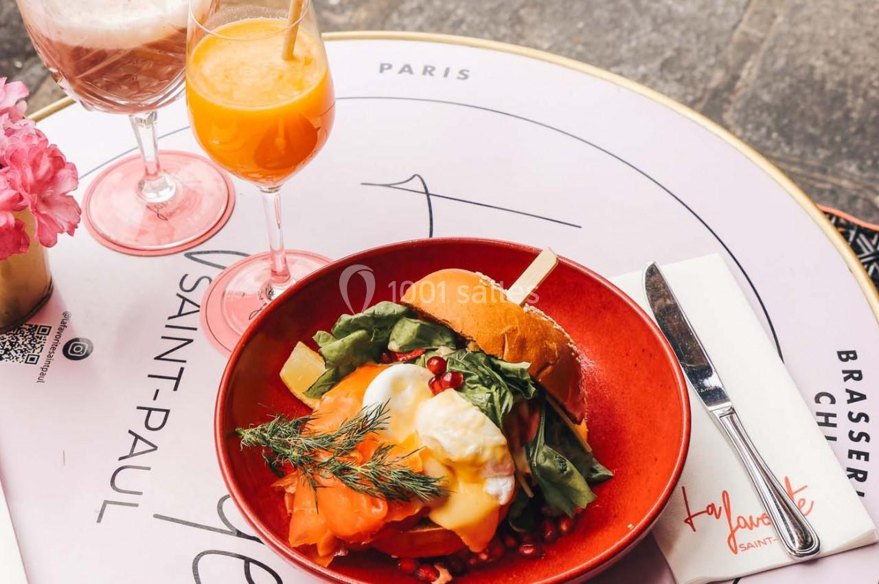 Assiette avec un burger garni de saumon, avocat et œuf poché, accompagnée de deux verres de jus sur une table de brasserie.