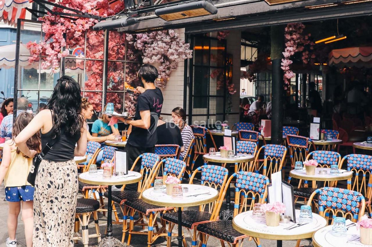 Terrasse d'un café avec des tables colorées, des clients assis et des serveurs en activité sous des décorations florales.
