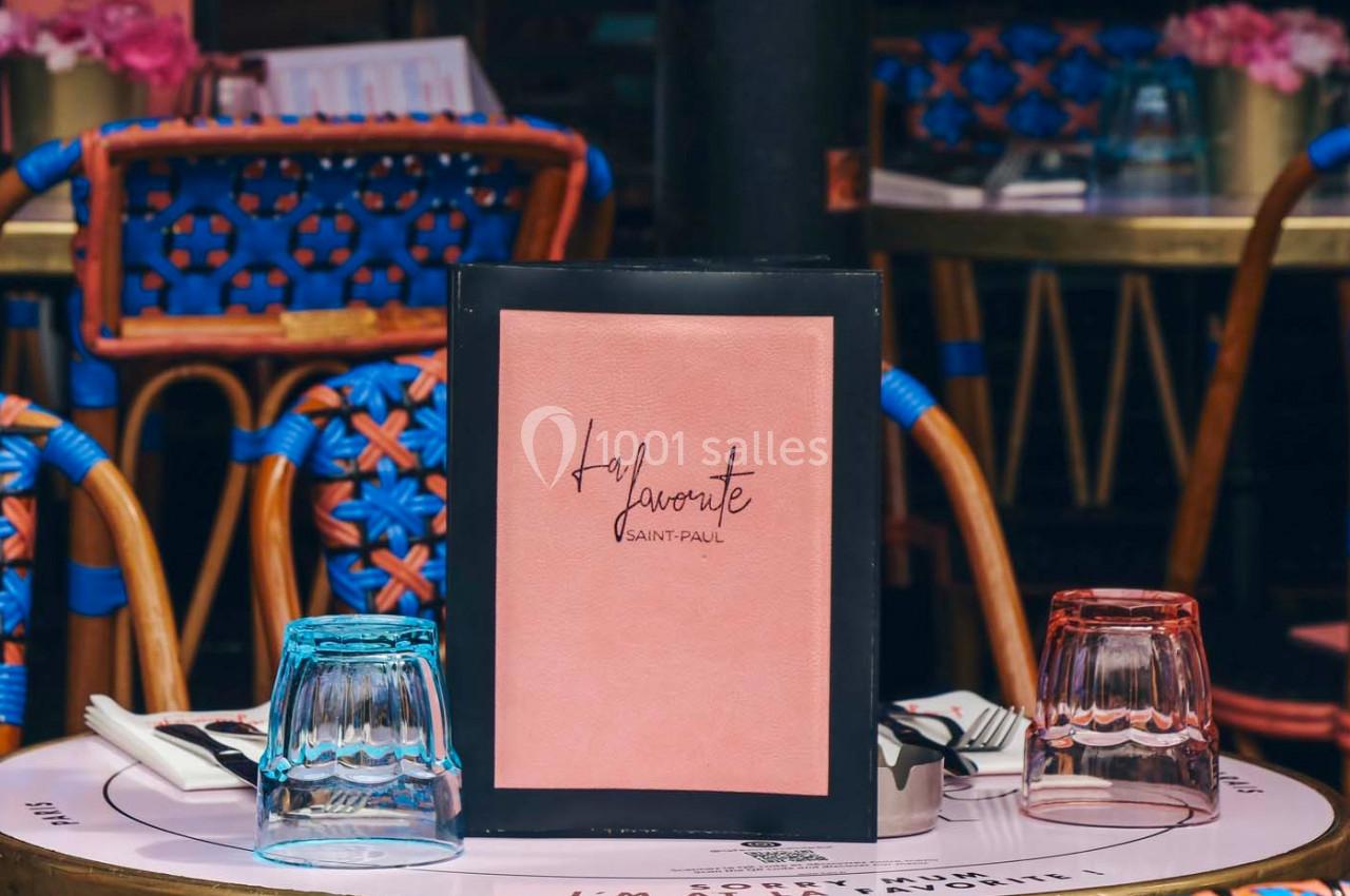 Menu rose encadré sur une table de restaurant avec verres colorés et chaises en osier en arrière-plan.