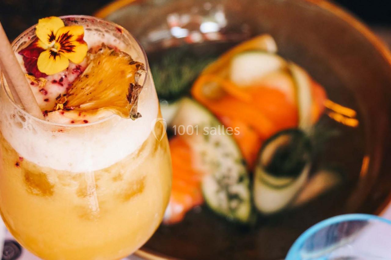 Verre de cocktail tropical garni d'une fleur et d'une tranche d'ananas, avec un plat de légumes en arrière-plan flou.