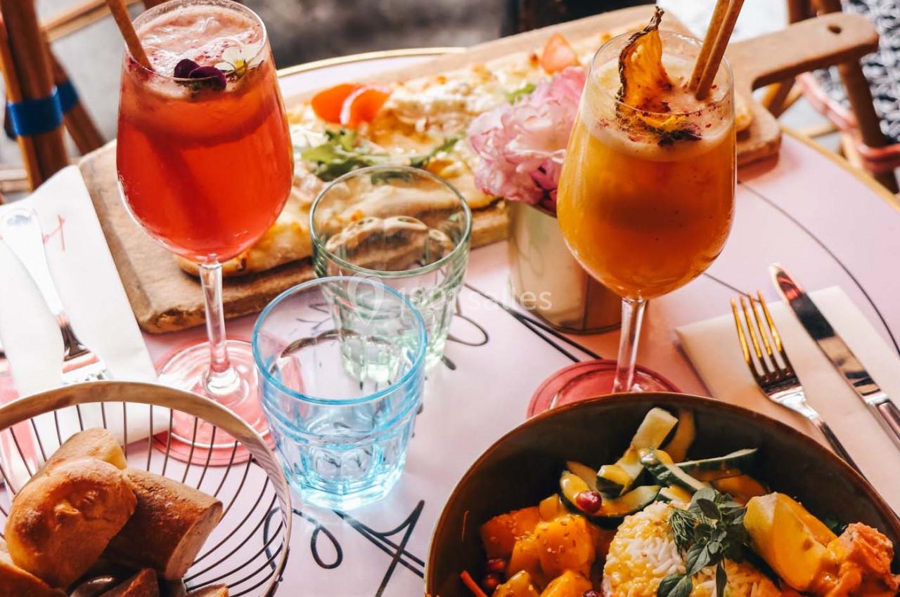 Deux cocktails colorés, une assiette de salade et une pizza sur une table dressée avec des verres et des couverts.