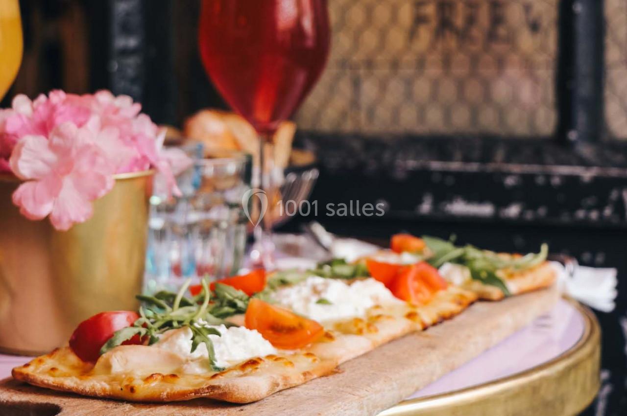 Pizza rectangulaire garnie de roquette, tomates et fromage, servie sur une planche en bois avec des boissons en arrière-plan.