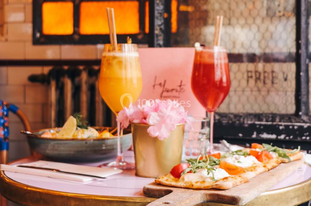 Boissons colorées, fleurs roses et plats de pizza sur une table dans un cadre chaleureux avec un four en arrière-plan.