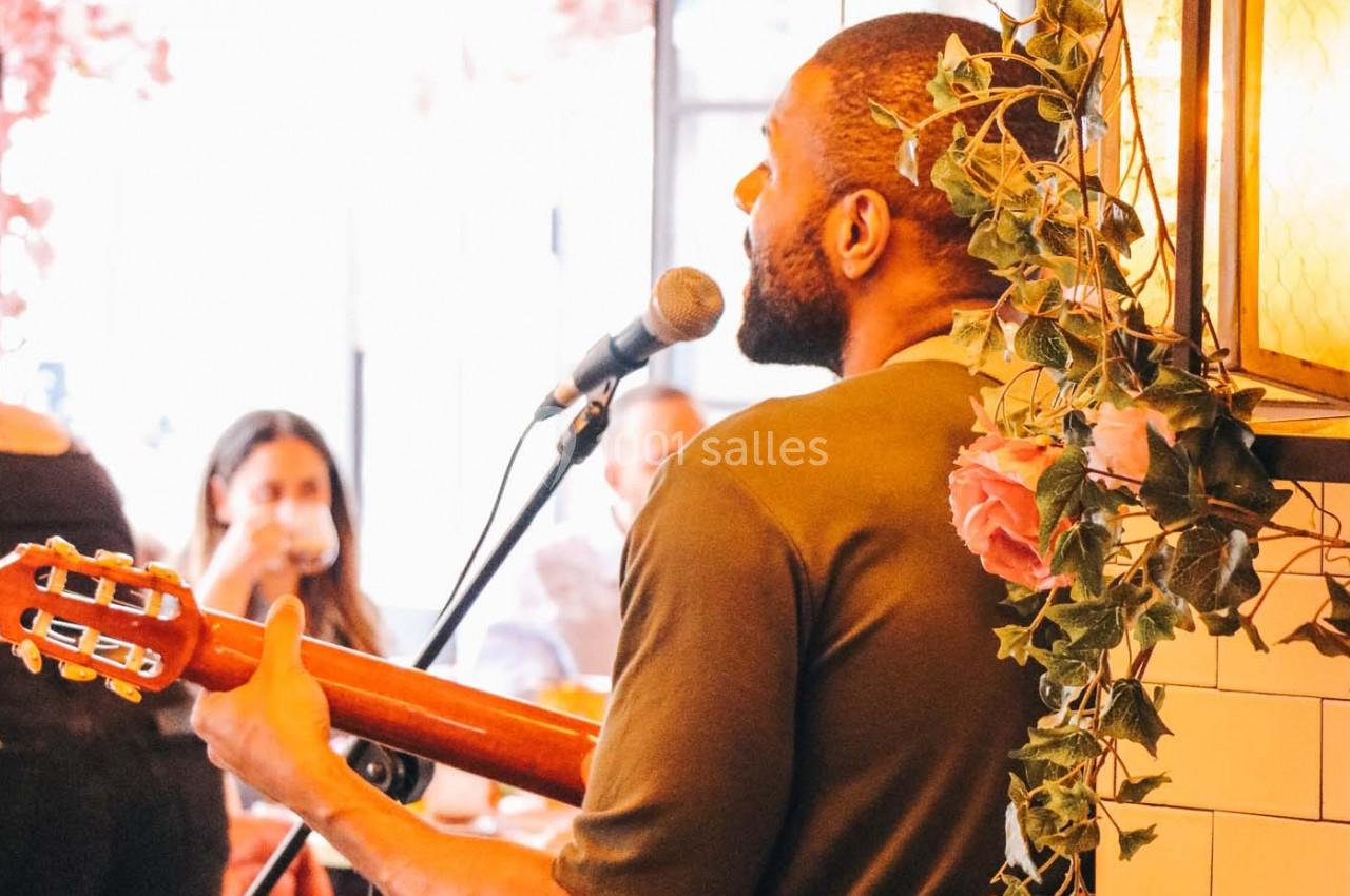 Un musicien joue de la guitare et chante au micro dans un café décoré de fleurs, devant un public attentif.
