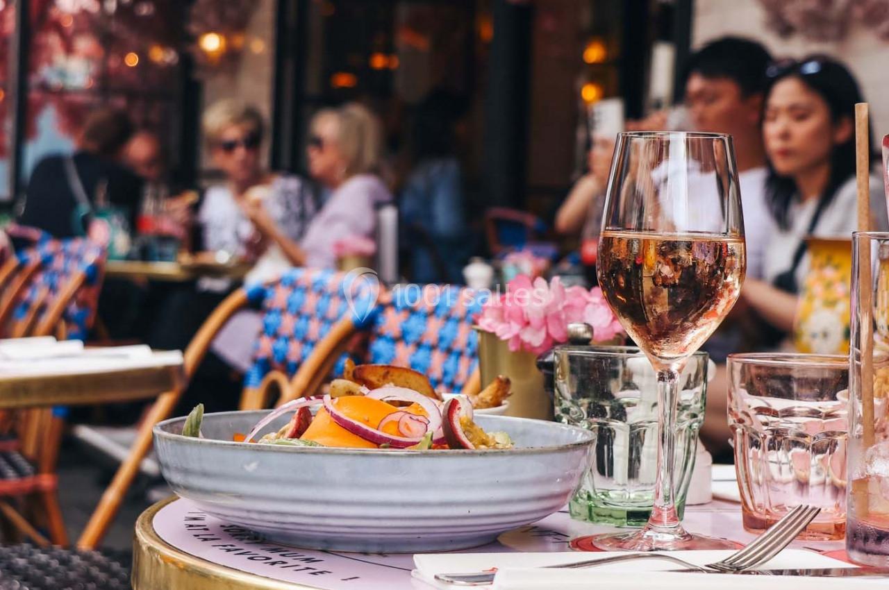 Assiette de salade garnie et verre de vin rosé sur une table en terrasse, avec des clients en arrière-plan flou.