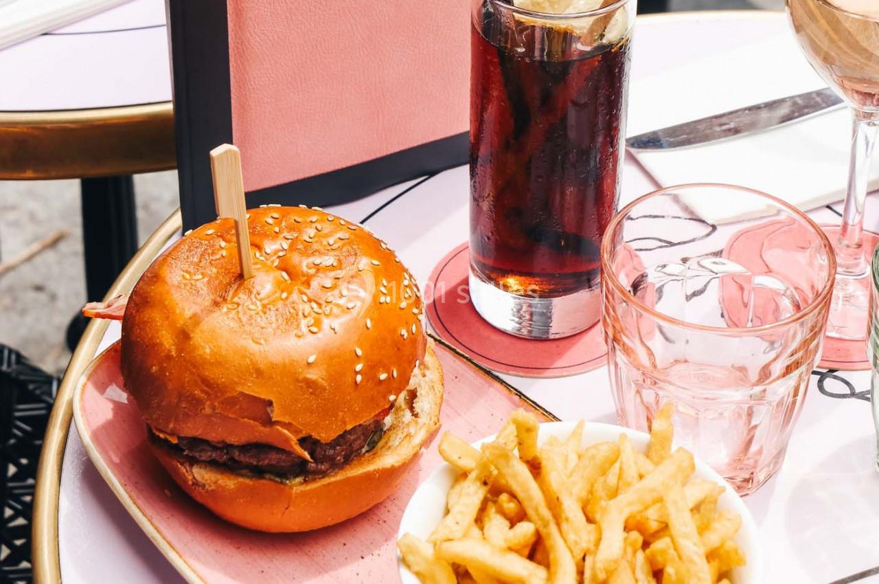 Burger avec pain au sésame, frites dorées, verre de soda et verre d'eau sur une table rose en terrasse.