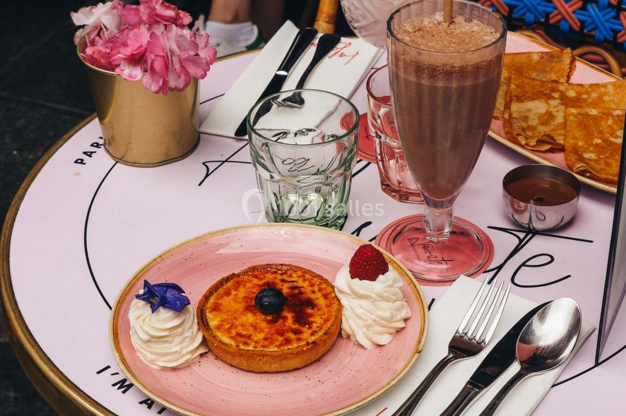 Une tartelette garnie de crème et fruits, accompagnée d'un milkshake chocolat, crêpes et couverts sur une table.
