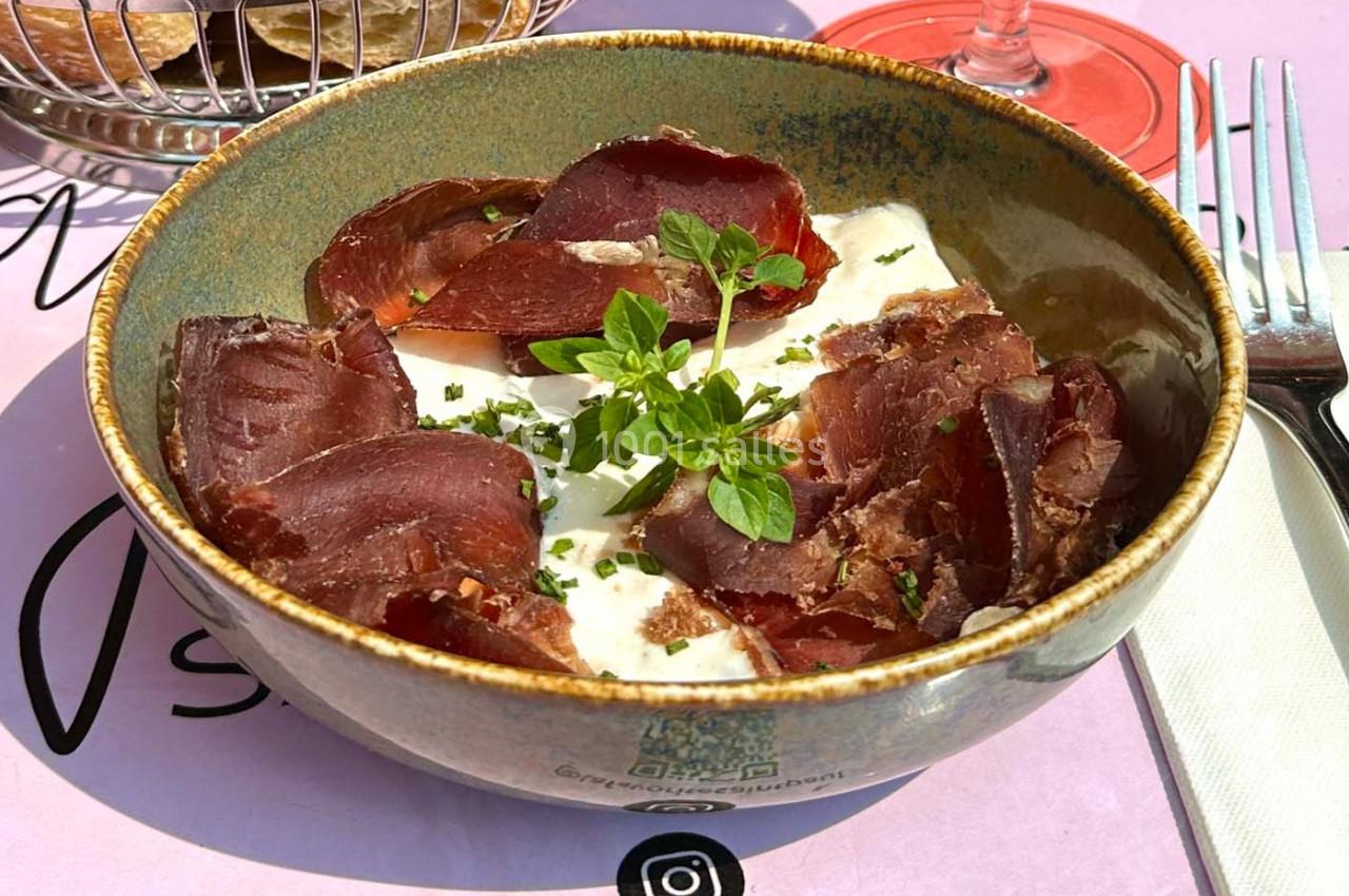 Bol contenant des tranches de viande séchée, une garniture d'herbes fraîches et une base crémeuse, posé sur une table rose.