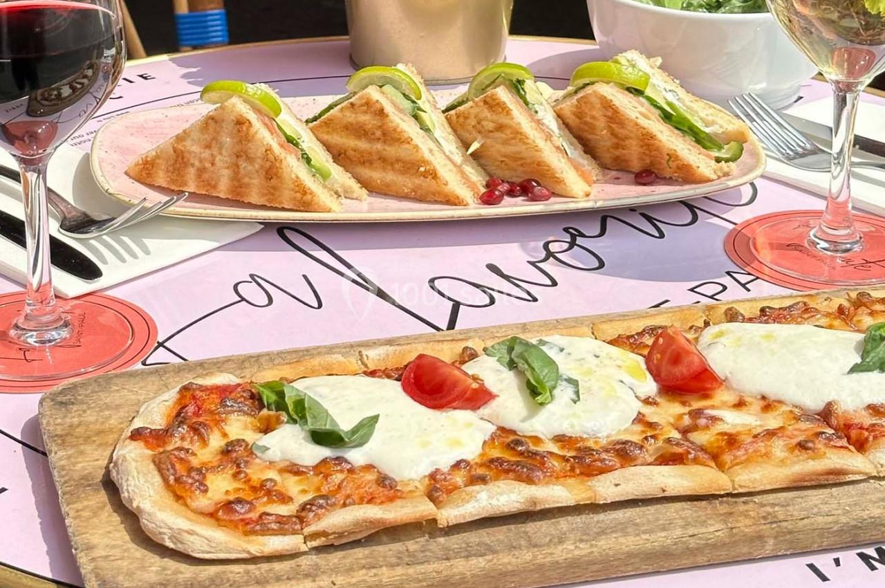 Assiette de sandwichs triangulaires et planche de pizza garnie de mozzarella et tomates, avec verres de vin rouge.