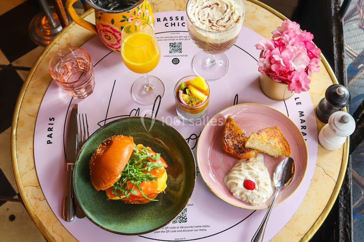 Table ronde avec un repas composé d'un burger, desserts, boissons et fleurs roses en décoration.