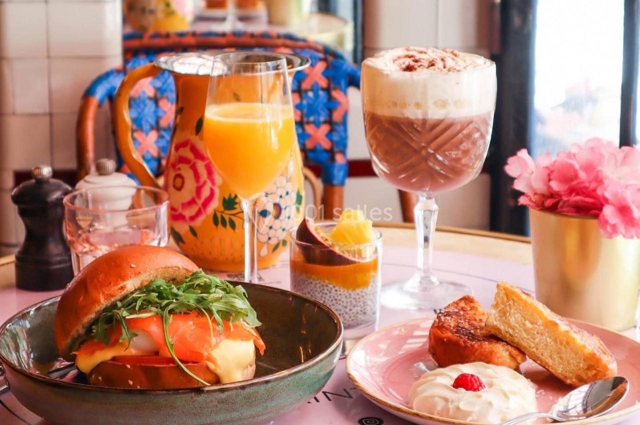 Petit-déjeuner composé d'un burger, d'un verre de jus, d'un chocolat chaud, de viennoiseries et de fruits sur une table…