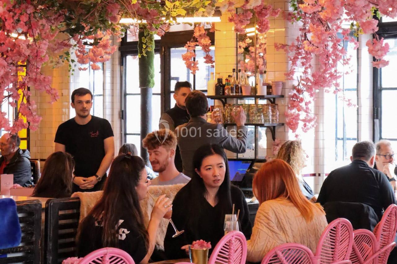 Salle de restaurant décorée de fleurs roses suspendues, avec des clients assis sur des chaises roses et noires.