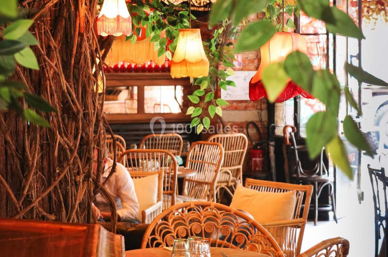 Intérieur d'un café avec des meubles en rotin, des lampes suspendues et une décoration végétale abondante.
