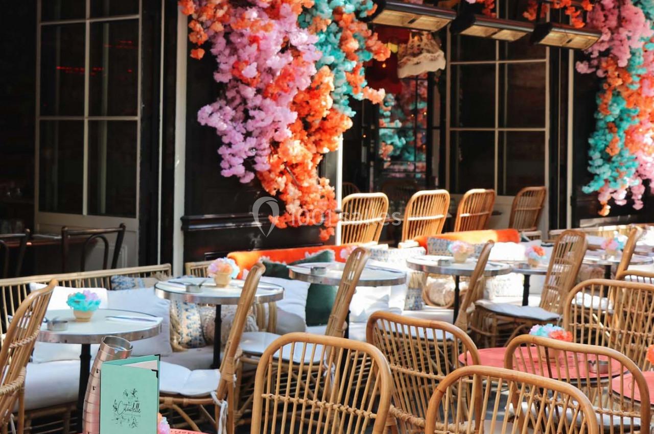 Terrasse d'un café avec des chaises en rotin, des tables rondes et des décorations florales colorées.