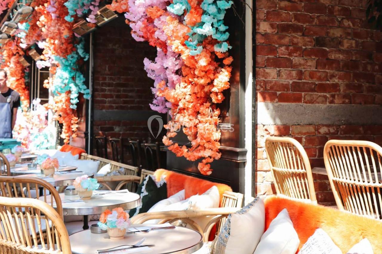 Terrasse ensoleillée avec tables, chaises en rotin et décorations florales colorées suspendues devant un mur de briques.