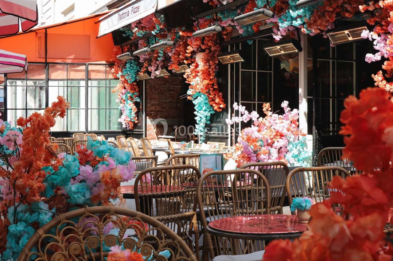 Terrasse de café décorée de fleurs colorées avec des chaises en rotin et des tables aux plateaux rouges.