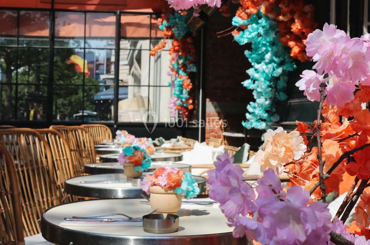 Terrasse de café décorée de fleurs colorées, avec des tables rondes et des chaises en rotin sous un auvent orange.
