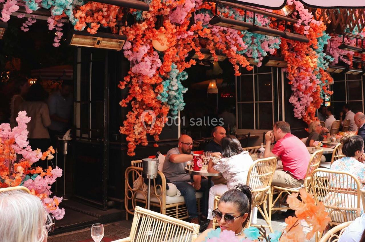Terrasse de café animée avec des clients assis et une décoration florale colorée en orange et rose.