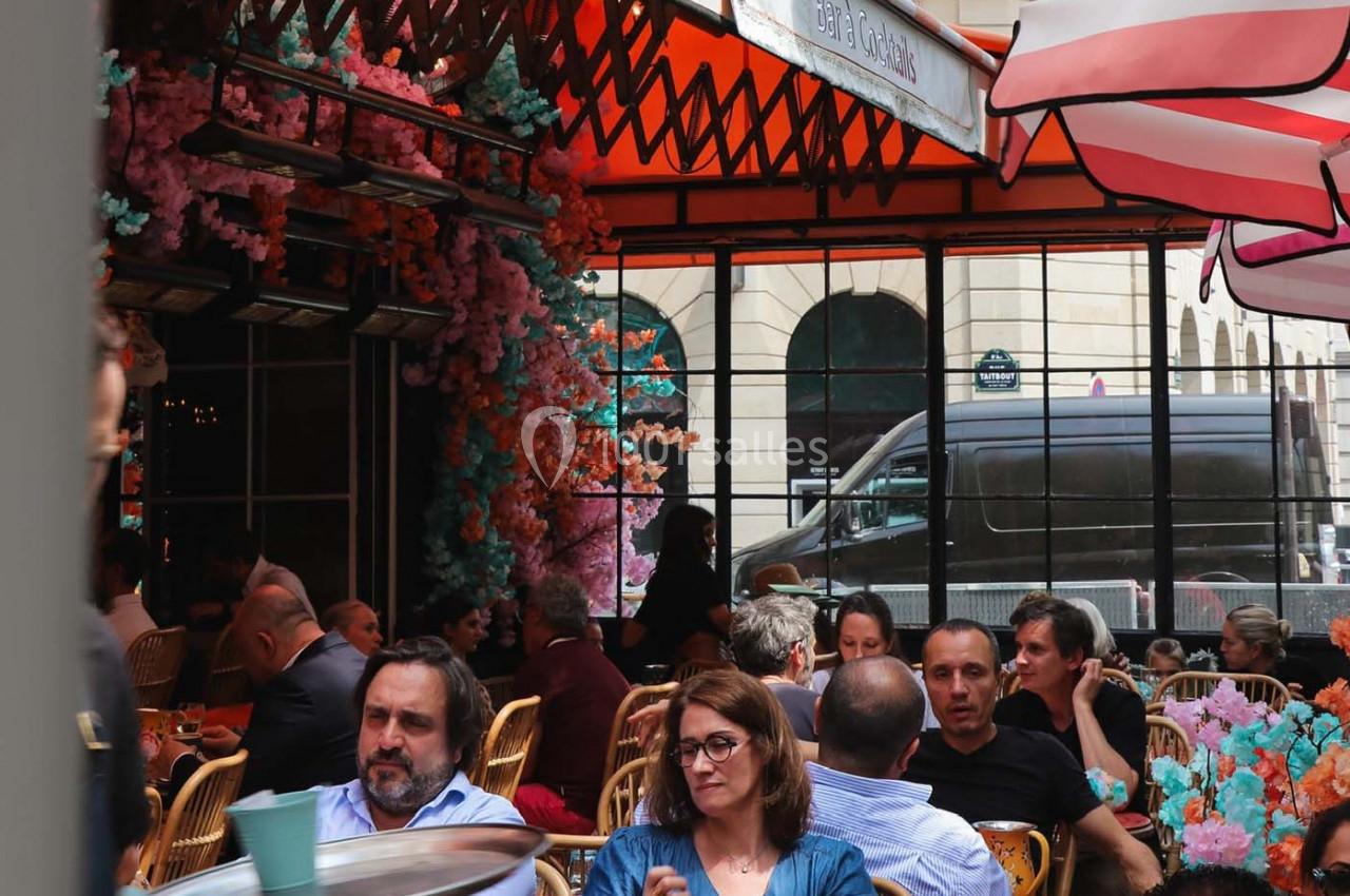 Terrasse d'un café animé avec des clients assis, décorée de fleurs colorées et protégée par des parasols.
