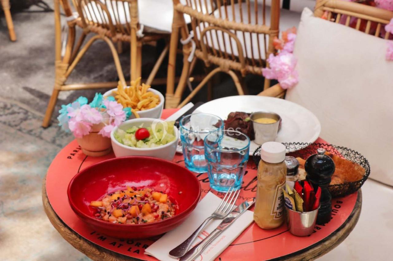 Table ronde avec des plats variés, dont une salade colorée, des frites, et un steak, sur une terrasse ensoleillée.
