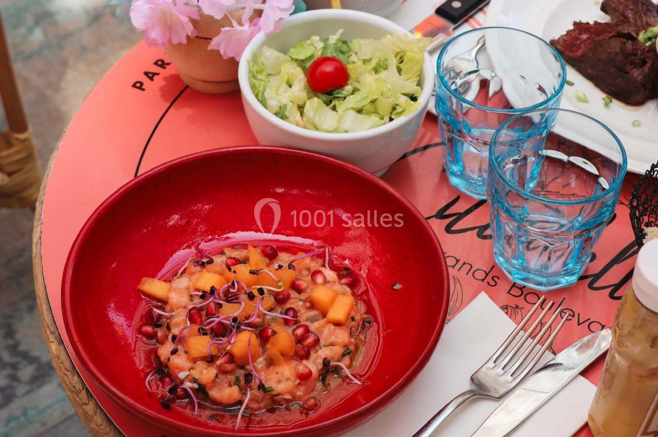 Assiette de tartare de saumon avec grenade et melon, accompagnée d'une salade verte et d'eau sur une table rouge.