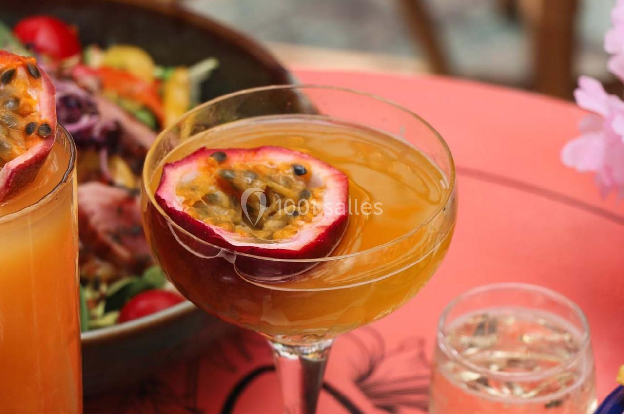 Verre de cocktail orange garni d'une demi-passion, posé sur une table rouge avec des plats en arrière-plan.