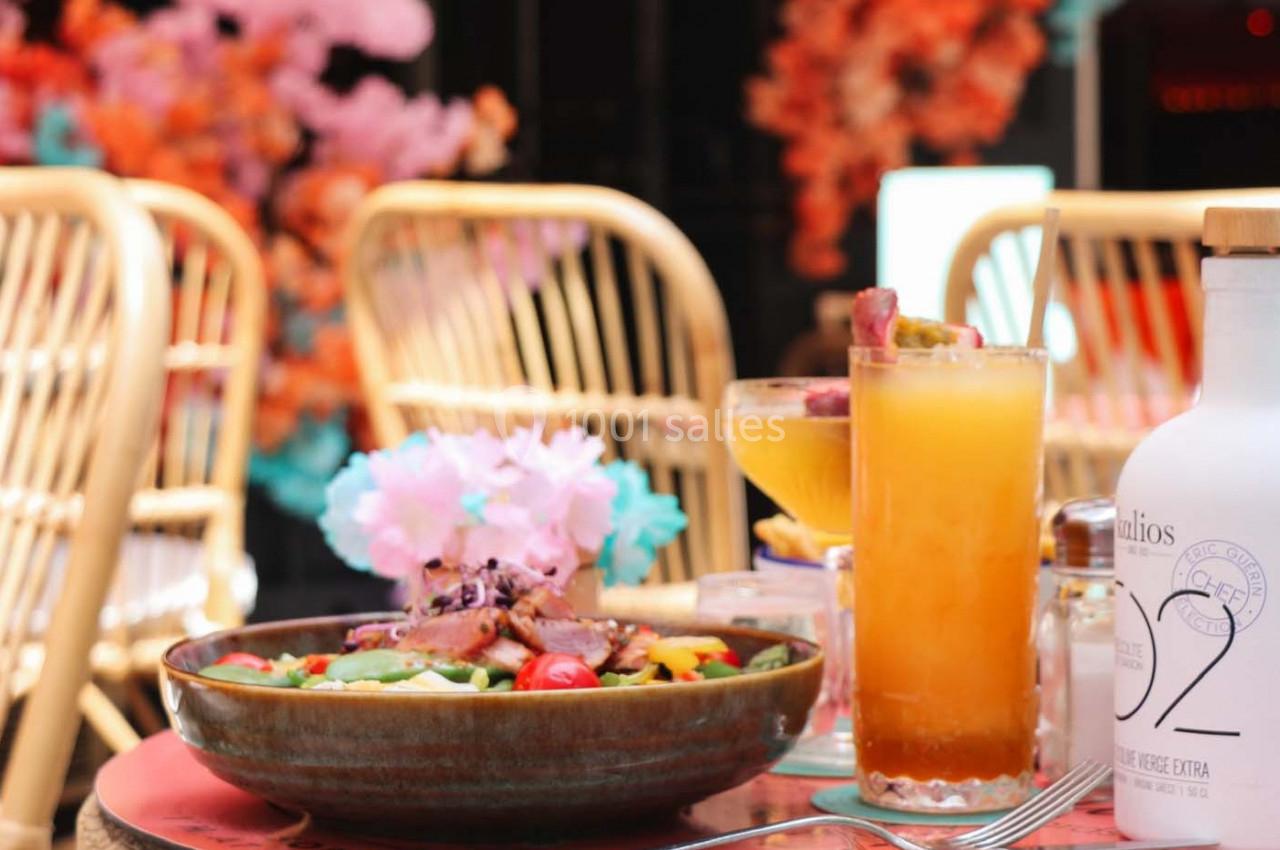 Assiette de salade colorée avec boissons fraîches sur une table en terrasse, décorée de fleurs et mobilier en rotin.