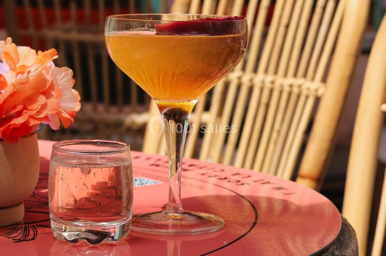 Verre à cocktail orange garni d'une tranche de fruit rouge, posé sur une table rose avec un verre d'eau et des fleurs.