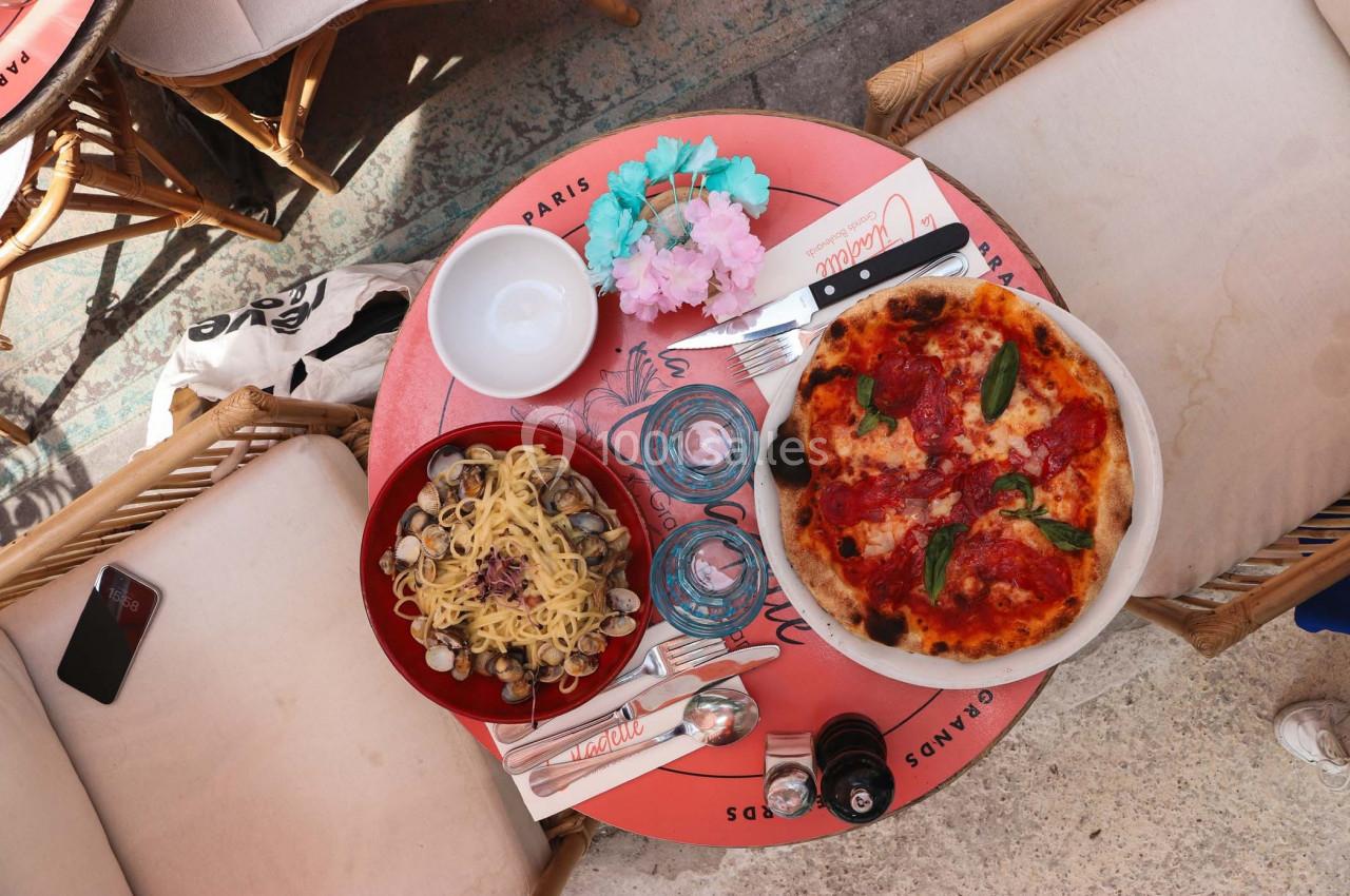 Table ronde avec une pizza margherita, un plat de pâtes aux palourdes, des verres, et des couverts dans un cadre extérieur.