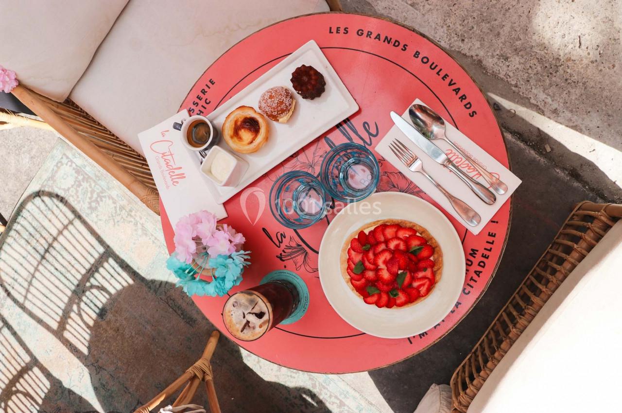 Table ronde avec une tarte aux fraises, viennoiseries, boissons et couverts, disposée en terrasse ensoleillée.