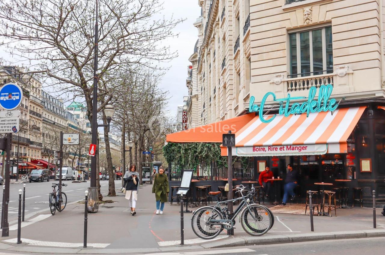 Terrasse d’un café parisien avec auvent orange et blanc, située à l’angle d’une rue bordée d’arbres et de bâtiments.