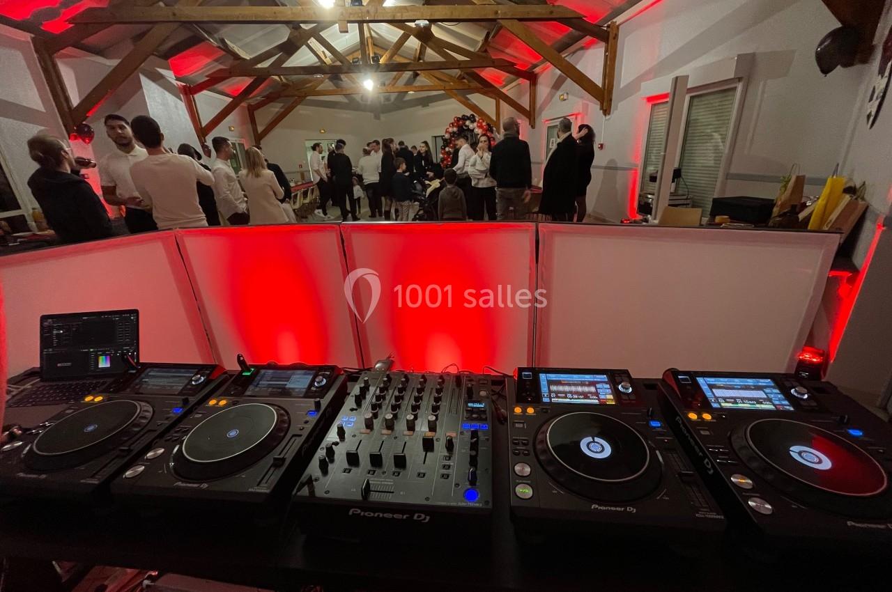 Vue depuis une table de mixage avec platines DJ, dans une salle éclairée en rouge où des invités discutent debout.