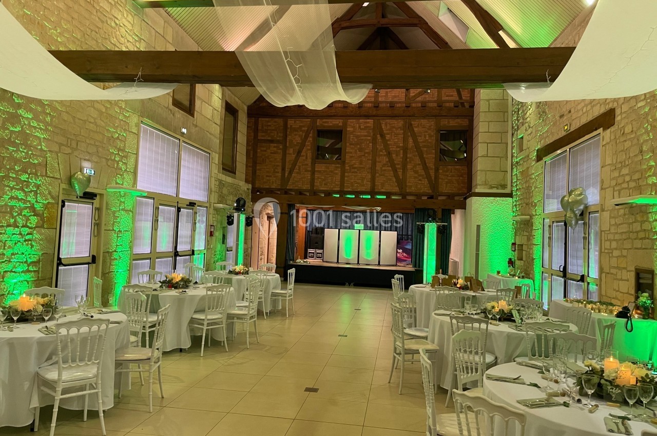 Salle de réception décorée avec des tables rondes, chaises blanches et éclairage vert, dans un cadre en pierre et bois.