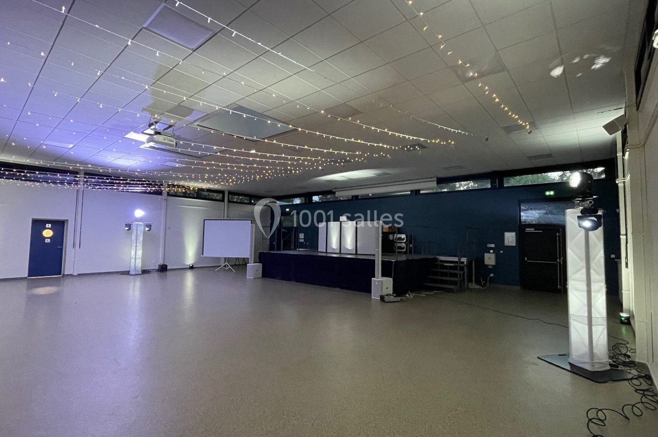 Salle vide avec scène, projecteurs, guirlandes lumineuses suspendues et écran de projection au fond.