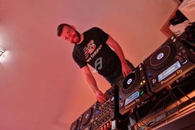 Un homme souriant mixe de la musique sur une table de mixage équipée de platines DJ dans une pièce éclairée en rouge.