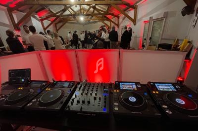 Vue depuis une table de mixage avec platines DJ, dans une salle éclairée en rouge où des invités discutent debout.