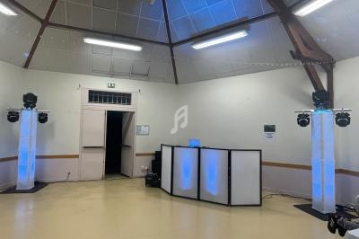 Salle avec un espace DJ équipé de lumières colorées et d'enceintes, dans un décor sobre avec plafond en bois.