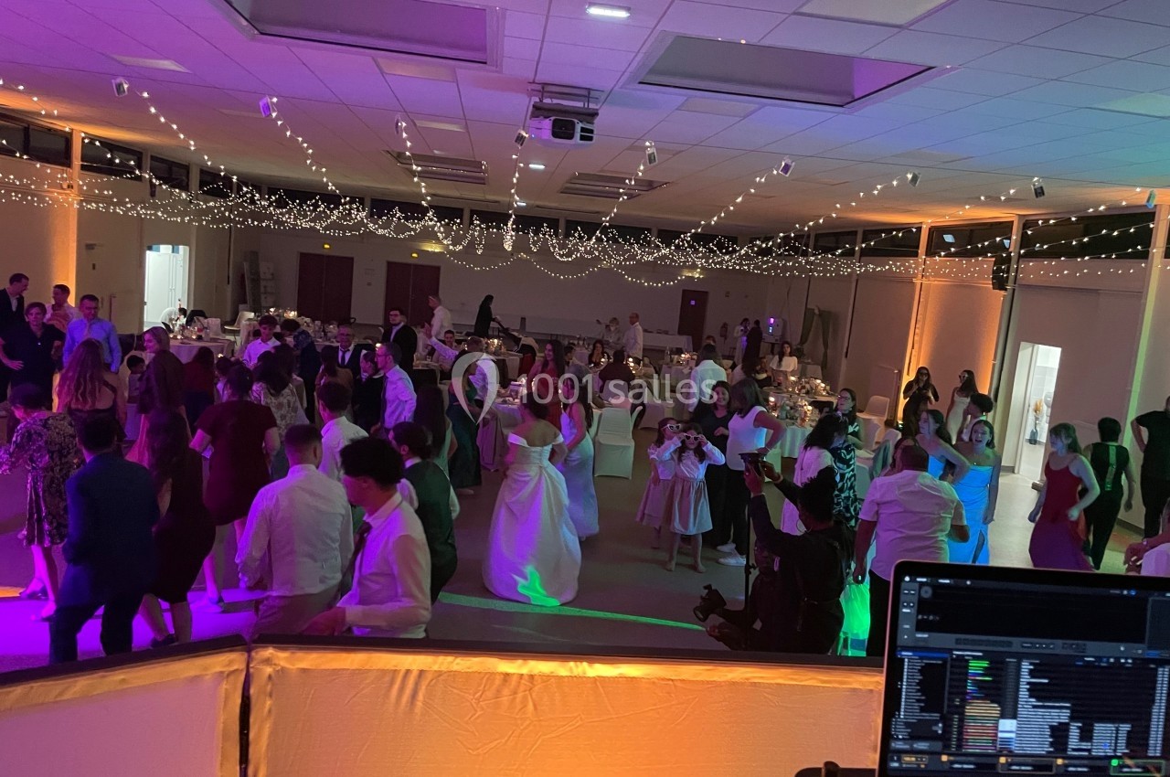 Salle de réception décorée avec des guirlandes lumineuses, des invités dansant et une console de DJ au premier plan.