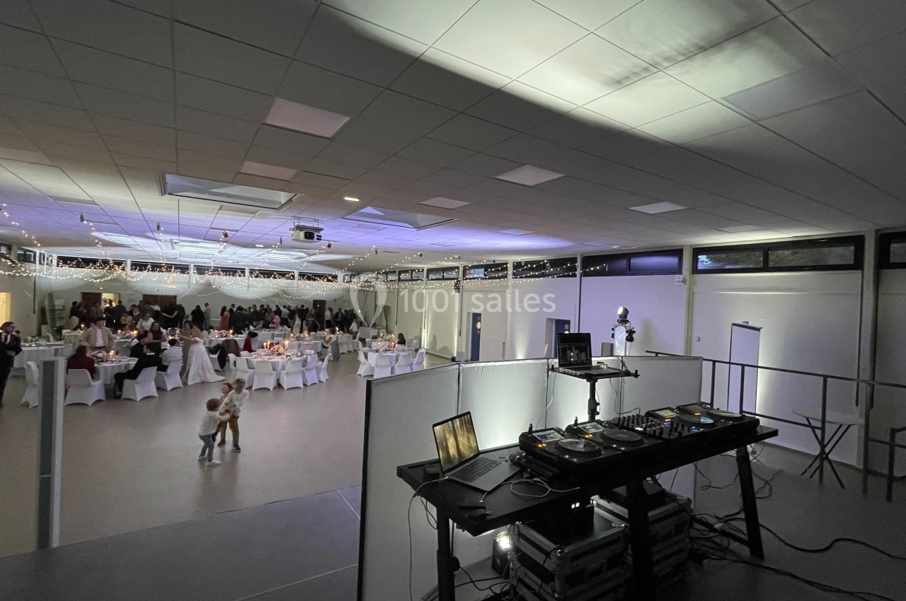 Salle de réception éclairée avec des invités assis à des tables décorées, vue depuis une table de mixage de DJ.