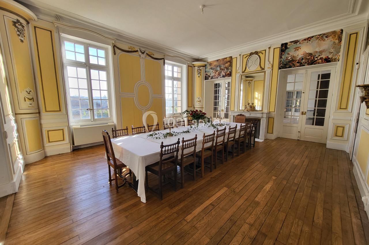 Salle à manger lumineuse avec parquet en bois, murs jaunes ornés, grande table dressée et fenêtres donnant sur l'extérieur.