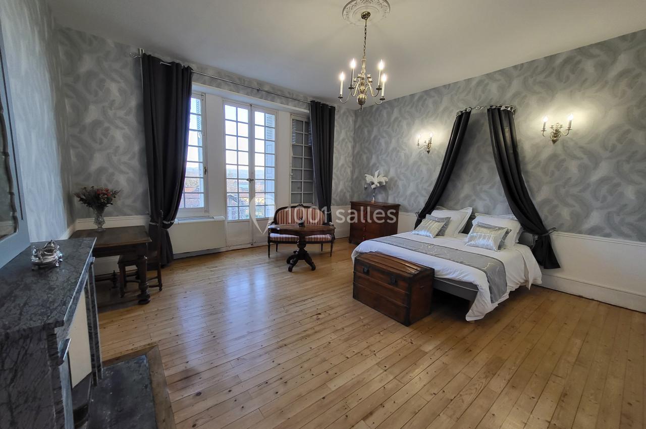 Chambre spacieuse avec parquet en bois, lit double, rideaux sombres, mobilier ancien et grande fenêtre donnant sur l…